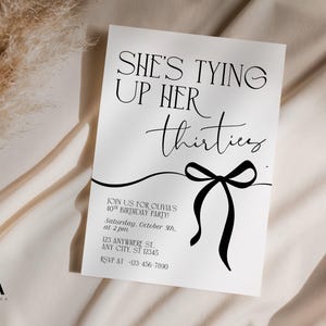 May include: A white invitation card with black text that reads "SHE'S TYING UP HER thirties". A black bow graphic is below the text. The invitation is for Olivia's 40th birthday party. The card is on a cream-colored fabric.