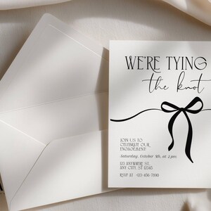 Puede incluir: Una tarjeta de invitaci&oacute;n de compromiso blanca con su sobre a juego. La tarjeta presenta el texto en negro WE'RE TYING the knot, seguido de los detalles del evento incluyendo fecha, hora y lugar, y un gr&aacute;fico negro de un lazo.