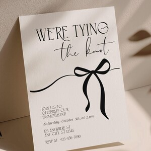 Puede incluir: Una tarjeta de invitaci&oacute;n de compromiso color crema con el texto negro WE'RE TYING the knot y un gr&aacute;fico de lazo negro. La tarjeta incluye los detalles: JOIN US TO CELEBRATE OUR ENGAGEMENT! Saturday, October 5th, at 2 pm, 123 ANYWHERE ST. ANY CITY, ST 12345, RSVP AT -123-456-7890.