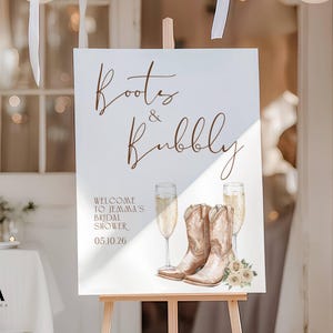 May include: A white rectangular sign on a wooden easel featuring the brown text Boots & Bubbly, Welcome to Jemma's Bridal Shower, and 05.10.26. The design includes illustrations of two champagne flutes, a pair of tan cowboy boots, and flowers.