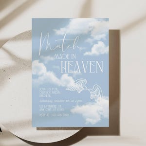May include: A rectangular bridal shower invitation card featuring a light blue sky background with white clouds. The text reads Match MADE IN HEAVEN, JOIN US FOR OLIVIA'S BRIDAL SHOWER, Saturday, October 5th, at 2 pm, 123 ANYWHERE ST, ANY CITY, ST 12345, RSVP AT 123-456-7890, and includes a white line drawing of angel wings.