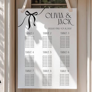May include: A white rectangular wedding seating chart sign with black text reading Olivia & Jack and Please Find Your Seat. The sign features a black line drawing of a bow at the top and lists nine tables with names printed in a black serif font.