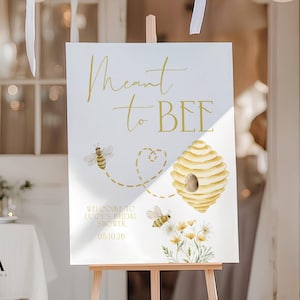 Może przedstawiać: Biały prostokątny znak na drewnianej sztaludze ze złotym napisem Meant to BEE, Welcome to Lucy's Bridal Shower oraz 05.10.26. Projekt zawiera akwarelowe ilustracje dwóch pszczół, ula oraz żółtych polnych kwiatów.