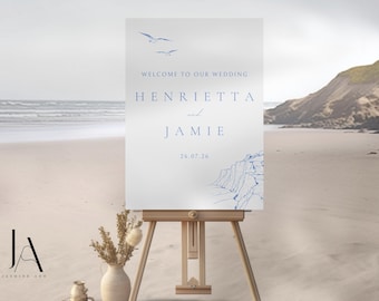 Op de afbeelding: Een rechthoekig welkomstbord voor een bruiloft op een houten ezel met blauwe tekst die Welcome to our wedding, Henrietta and Jamie en de datum 26.07.26 vermeldt. Het ontwerp bevat blauwe lijntekeningen van twee meeuwen en kustkliffen.