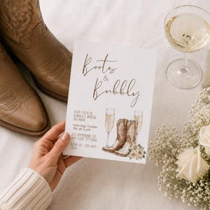 Puede incluir: Una tarjeta de invitación rectangular blanca para una despedida de soltera con el texto Boots & Bubbly en letra cursiva marrón. La tarjeta presenta ilustraciones de dos copas de champán, un par de botas vaqueras marrones y un ramo de rosas blancas.