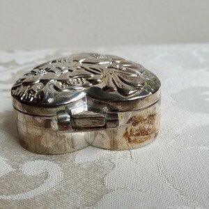 Puede incluir: Una caja de coraz&oacute;n de plata con un dise&ntilde;o floral en la tapa. La caja est&aacute; cerrada y la tapa est&aacute; ligeramente entreabierta.