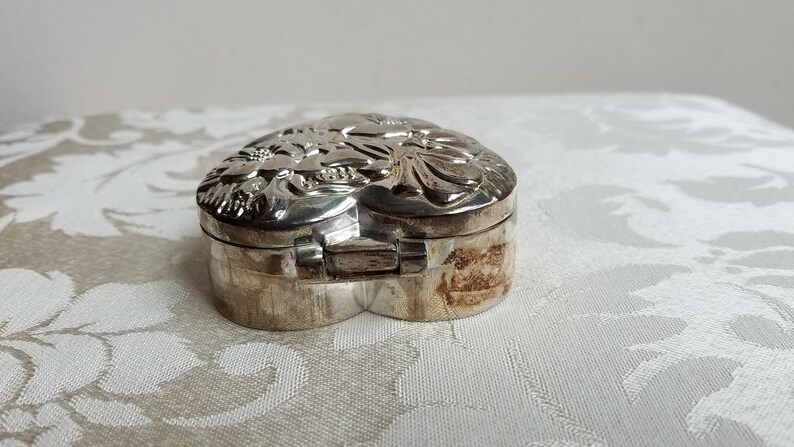 Puede incluir: Una caja de coraz&oacute;n de plata con un dise&ntilde;o floral en la tapa. La caja est&aacute; cerrada y la tapa est&aacute; ligeramente entreabierta.