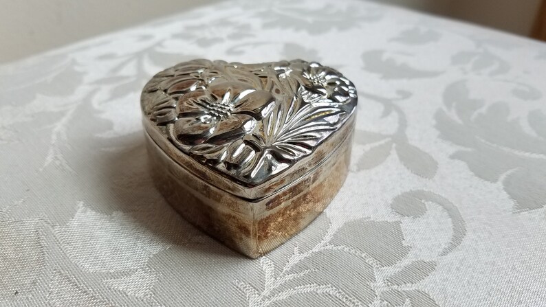 Puede incluir: Una caja de coraz&oacute;n de plata con un dise&ntilde;o floral en relieve.
