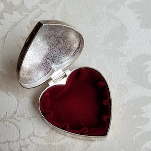 Puede incluir: Una caja de coraz&oacute;n de plata con un interior de terciopelo rojo. La caja est&aacute; abierta y la tapa est&aacute; sobre la mesa.