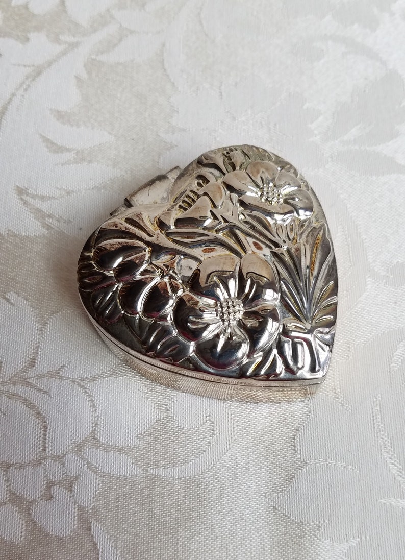 Puede incluir: Una caja de coraz&oacute;n de plata con un dise&ntilde;o floral. La caja est&aacute; cerrada y tiene una tapa con bisagras.