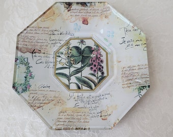 Puede incluir: Un plato de vidrio octogonal blanco con un diseño floral y texto escrito a mano en francés. El plato presenta una ilustración botánica de una planta con flores rosas y hojas verdes. El texto incluye frases como "Gloire" y "Je vais me et me".