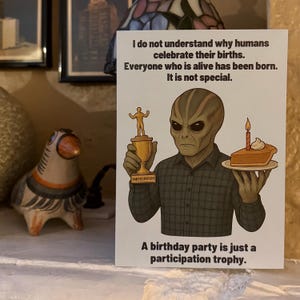 May include: A rectangular greeting card featuring a gray alien in a dark plaid shirt. The alien holds a gold participation trophy and a slice of cake with a candle. Text reads: I do not understand why humans celebrate their births. Everyone who is alive has been born. It is not special. A birthday party is just a participation trophy.