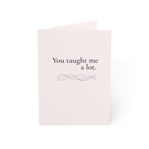 May include: A folded greeting card with a pale pink cover. The card features the text "You taught me a lot" in a dark grey font, with a decorative wavy line design underneath. The card is blank inside.