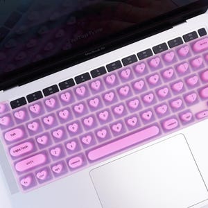 Könnte beinhalten: Eine rosa herzförmige Tastaturabdeckung auf einem silbernen Laptop. Die Abdeckung hat herzförmige Tasten und eine passende rosa Leertaste. Der Laptop-Bildschirm ist im Hintergrund sichtbar.