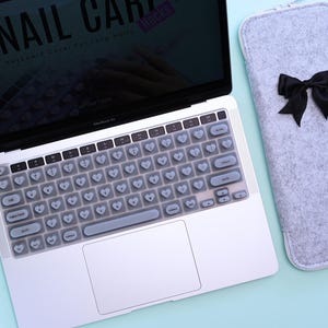 Puede incluir: Un ordenador portátil MacBook Air plateado con una funda de teclado en forma de corazón y una funda de fieltro gris con un lazo negro. La pantalla muestra el texto "NAIL CARE Keyboard Cover for Long Nails".