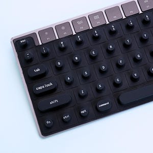 May include: A close-up of a black and silver computer keyboard. The keyboard has black keys with white lettering and a silver frame. The keys include letters, numbers, and function keys. The keyboard is on a light blue surface.