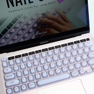 May include: A white MacBook Air laptop with a keyboard cover. The cover has rounded keys and is designed to protect the keyboard. The screen displays text that reads "Keyboard Cover For Long Nails" and "TapTapType".