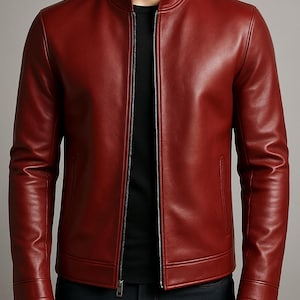 Puede incluir: Chaqueta de cuero roja con cuello alzado y cremallera completa. La chaqueta tiene dos bolsillos laterales y mangas largas. La persona lleva una camiseta negra y vaqueros azul oscuro.