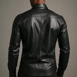 May include: A black leather shirt with long sleeves and a button-down collar. The shirt has a tailored fit and a classic design. The model is wearing black trousers. The background is a solid grey.