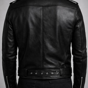 May include: Black leather jacket with a classic design. Features include a notched collar, shoulder epaulets, and a belt at the waist. The jacket has zippered pockets and cuffs. The model is wearing black jeans.