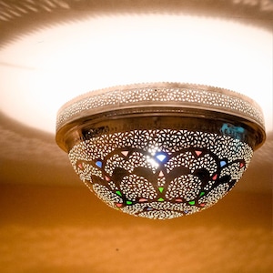 May include: Ornate, antique-style ceiling lamp with a perforated metal shade. The lamp's design features intricate cut-out patterns and colorful glass inserts. The light casts decorative shadows on the ceiling and surrounding wall.