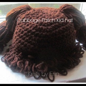 May include: A dark brown crocheted hat featuring yarn pigtails on the sides and a curly fringe along the bottom edge. The text Cabbage Patch Kid hat and Knits by Erin is printed on the front of the dark brown yarn construction.