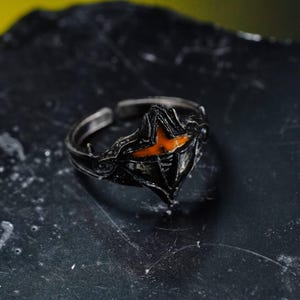 May include: A black adjustable ring with an orange lightning bolt design. The ring has a detailed, textured appearance and is set against a dark background. The ring's design is centered, showcasing its unique shape and color contrast.