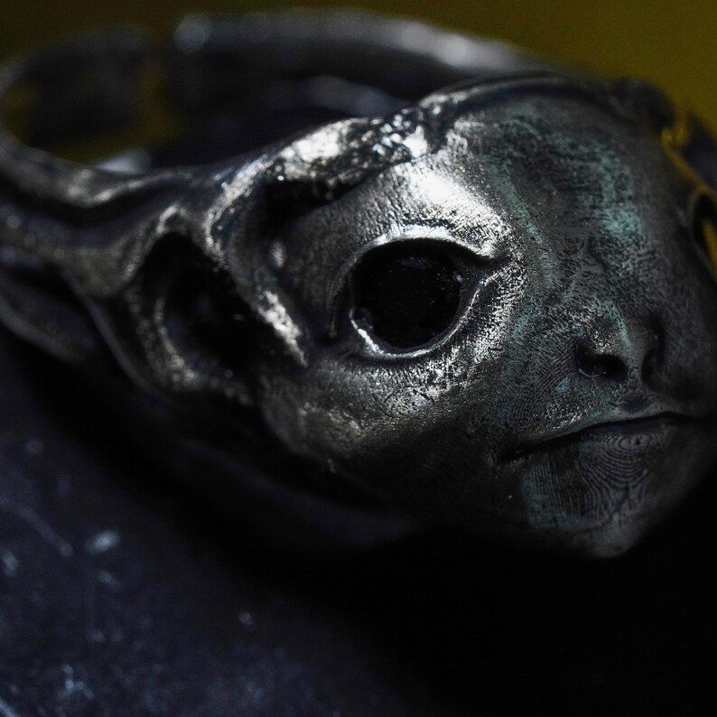 May include: A dark silver-toned ring featuring a sculpted creature head with deep, recessed eye sockets and a textured, weathered finish. The metal surface displays a metallic sheen with subtle dark oxidation, creating a detailed, three-dimensional form.