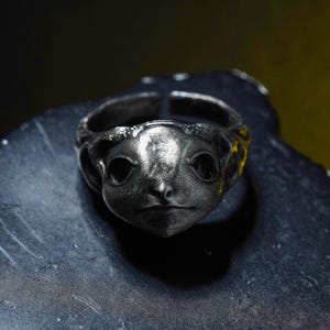 May include: A dark silver-toned metal ring featuring a sculpted creature face with large, smooth, black inset eyes, a pointed nose, and a small mouth. The band is open at the back, showing a textured, weathered finish across the entire surface.