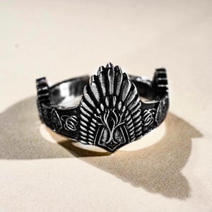 May include: A silver-toned ring with an ornate design. The ring features a central tree motif with a feather-like design above it. The band has detailed patterns, and the ring has a vintage or fantasy aesthetic.