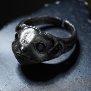 May include: A dark, metallic ring featuring a sculpted creature head with prominent, smooth eyes and a textured, weathered surface. The band is open at the back, showing a continuous, rugged design that wraps around the finger with a dark finish.