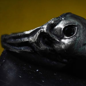May include: A silver-toned metal ring featuring a sculpted animal skull design with a textured, weathered finish. The eye socket of the skull is set with a single, small, round, faceted black gemstone, creating a dark and detailed appearance.