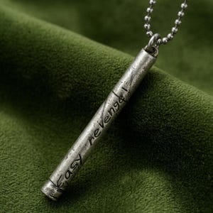 May include: A silver-toned cylindrical pendant necklace with the words "Easy revenge!" etched on the side. The pendant hangs from a ball chain. The background is a textured, dark green fabric.