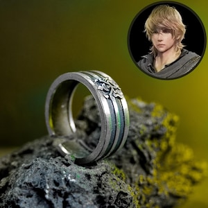 May include: Silver ring with a detailed design. The ring features vertical stripes in silver, black, and green. A small, circular portrait of a person with blonde hair is in the background.