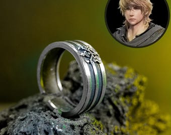 May include: Silver ring with a detailed design. The ring features a series of vertical stripes in silver, black, and green. A small, circular portrait of a person with blonde hair is in the background.
