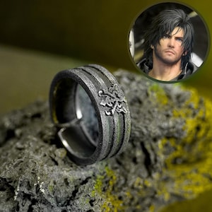May include: A silver ring with a textured band and floral detailing. The adjustable ring is displayed on a textured rock. A circular image of a man with dark hair is in the background.