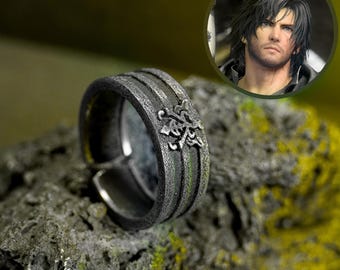 May include: A silver-toned ring with a textured band and floral design. The ring is adjustable and sits on a textured rock surface. A circular image of a man with dark hair is in the background.