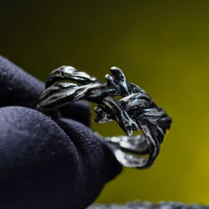 May include: A silver-toned ring with a textured, intertwined branch design. The ring's surface has a dark, oxidized finish, enhancing the intricate details of the branches. The ring is set against a dark background with a yellow-green gradient.