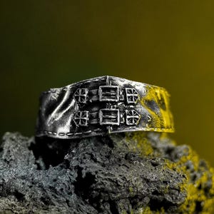 May include: A silver ring with a textured band and buckle-style embellishments. The ring has four decorative elements that look like buckles and cross motifs. The ring is shown on a dark, textured surface.