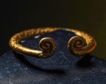 May include: A gold-colored cuff bracelet with a textured surface and spiral ends. The open-ended design features intricate detailing and a warm, metallic finish. The bracelet is displayed on a dark, textured surface.