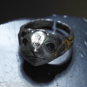 May include: A silver-toned metal ring featuring a sculpted creature face with prominent dark eyes and a textured band. The design includes an open back construction, showcasing a weathered, metallic finish with detailed facial features and contours.