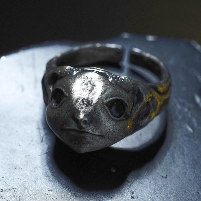 May include: A silver-toned metal ring featuring a sculpted creature face with prominent dark eyes and a textured band. The design includes an open back construction, showcasing a weathered, metallic finish with detailed facial features and contours.