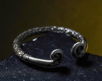 May include: A silver-colored open cuff bracelet with a textured surface and decorative details. The bracelet features curled ends, resting on a dark, textured surface. The design is intricate and detailed.