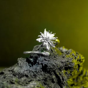 May include: A silver ring featuring a detailed flower design. The ring sits on a dark, textured surface with green moss. The flower has multiple petals and is the focal point of the jewellery.