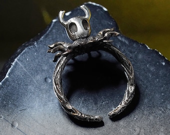 May include: A silver-colored ring featuring a detailed design of a character's head with horns, set atop a textured band. The ring's band has an open design. The ring is likely made of metal.