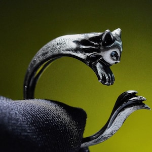 May include: A silver-toned ring featuring a stylized cat design. The cat's head and paws form the top of the ring, with the tail curving around the finger. The cat's face has white accents, and the ring is set against a green background.