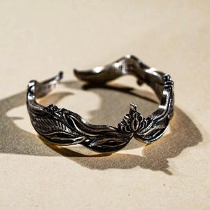 May include: A silver-toned adjustable ring with an ornate leaf and floral design. The ring features detailed carvings of leaves and a central floral element. The open design allows for size adjustment. The ring is displayed on a neutral background.