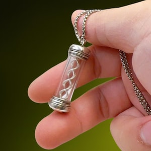 May include: A clear glass pendant necklace with a silver-colored chain. The pendant is a small, cylindrical vial containing a silver-colored, double-helix design. The vial has silver-colored caps on each end.