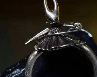 May include: A silver-toned ring featuring a stylized figure with a horned head, a pleated skirt, and a sword. The figure is secured to the ring with a safety pin. The ring is set against a dark background.
