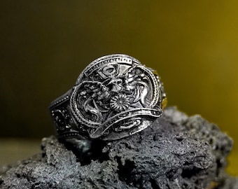 May include: A detailed silver ring featuring a central design with two mythical creatures flanking a floral element. The ring has intricate patterns and is set against a dark, textured background, showcasing its ornate details.